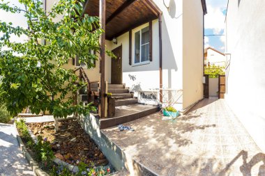 Veranda, merdivenler ve evin arka bahçesindeki kulübeye giriş. Girişteki tepe örtüsü. Ayakkabılar verandada. Ağaçlı arka bahçe. Güneşli bir gün.