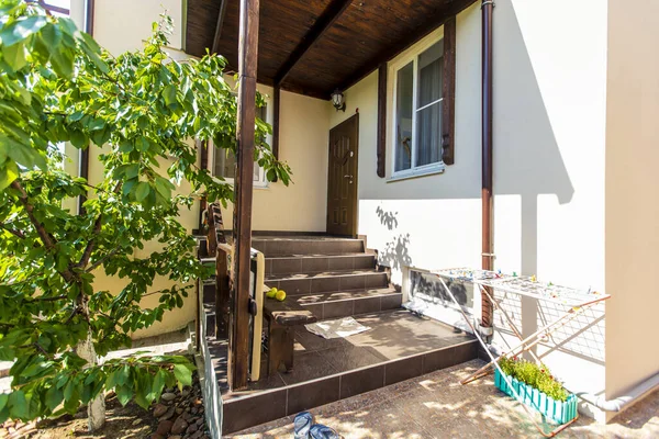 Portico e porta d'ingresso al cottage. Baldacchino di legno, alberi verdi e foglie in una giornata di sole. Scarpe all'ingresso — Foto Stock