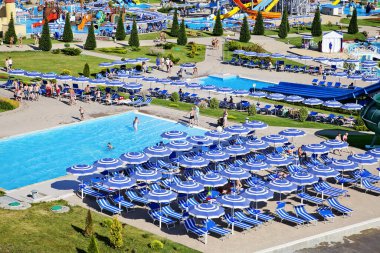 Güneşli bir günde su parkının merkez sokağı. Suyun etrafında kayar ve biner. Şemsiyeler ve güneş yatakları havuzun kenarında.