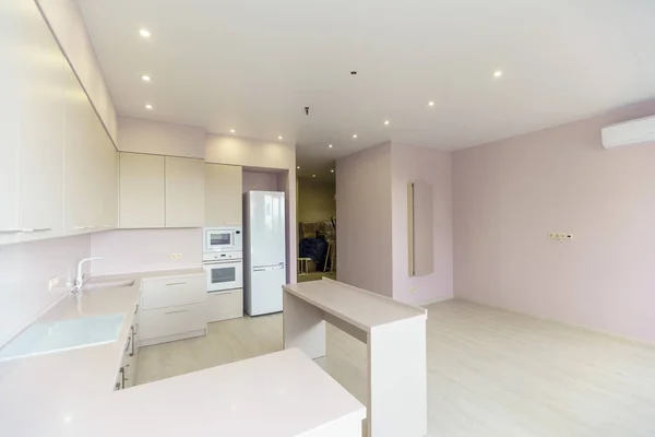 Велика кімната з рожевими стінами і білим кухонним гарнітуром. Кухонні меблі нові з усією кухонною технікою. Перед кухнею білий стіл. Свіжий, новий ремонт . — стокове фото