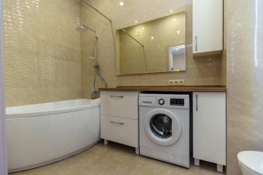 Kahverengi lavabosu ve tezgahı olan banyo, beyaz dolaplar, büyük ayna, çamaşır makinesi, tuvalet. Bej duvar kiremitleri