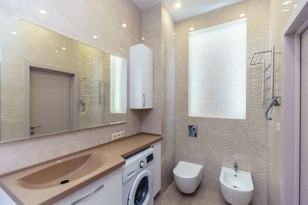 Ванная комната с коричневой раковиной и столешницей, белые шкафы, большое зеркало, стиральная машина, туалет. Бежевая плитка — стоковое фото
