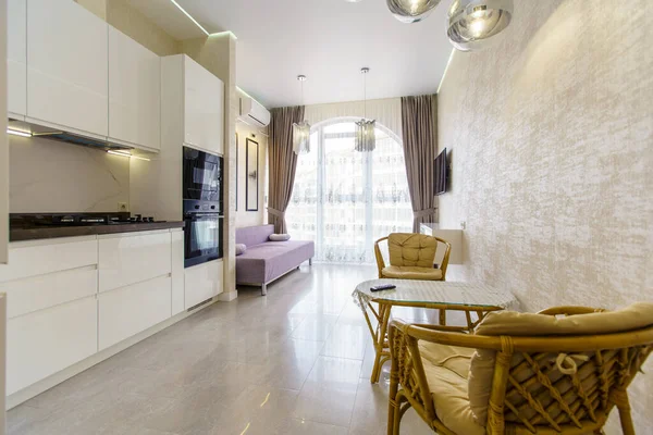 Kitchen room with a fresh renovation in beige tones. White kitchen set with kitchen appliances. semicircular window with curtains. TV, sofa and wicker furniture-table and two chairs