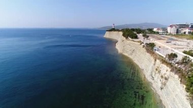 Karadeniz kıyısı boyunca İHA uçuşu. Yüksek kayalık uçurum. Aşağıda bir kiremit plajı şeridi var. Uzakta, Gelendzhik deniz feneri. 