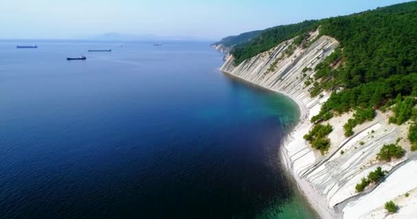 Voo sobre a costa do mar negro de Gelendzhik para Novorossiysk. O drone voa sobre altas rochas inclinadas. As montanhas estão cobertas de pinheiros. À distância Novorossiysk e navios no — Vídeo de Stock