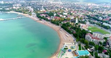 Gelendzhik tatil köyünün merkez plajı kuş bakışı manzaralı. Sıralar dolusu güneş şezlongları ve kumdaki şemsiyeler. İnsanlar yüzer ve güneşlenir. Zümrüt suyu. Sahil boyunca bir kiriş seti var.