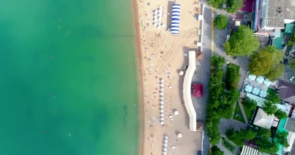鳥の目のビューからゲレンツィクリゾートの中央のビーチ。サンラウンジャーや傘の列で砂浜を飛んでいます。人々は泳いで日光浴をします。ゲレンツィクの美しいエメラルドグリーンの水 — ストック動画