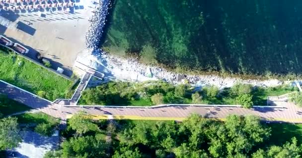 Promenada i żwirowa plaża, widok z góry. Długie cienie z latarni i ludzie ze słońca o zachodzie słońca. Szmaragdowe morze. Leżaki i parasole. Uzdrowisko Gelendzhik, gruby przylądek. Ludzie chodzą wzdłuż — Wideo stockowe