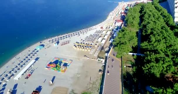 Stranden på orten staden Gelendzhik i området Tolstoy Cape. Videofilmning från en drönare. Det finns rader av solstolar och halmparasoller på den steniga stranden. Grönt träd. Banvallen med — Stockvideo