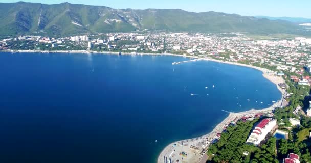 Gelendzhik-öböl madártávlatból. M strand esernyőkkel és napozóágyakkal a tenger mentén. Felszerelés korláttal. Jachtok és hajók a tengeren. Hegyek a háttérben. — Stock videók