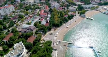 Gelendzhik tatil köyünün merkez plajı kuş bakışı manzaralı. Sıralar dolusu güneş şezlongları ve kumdaki şemsiyeler. İnsanlar yüzer ve güneşlenir. Zümrüt suyu. Sahil boyunca bir kiriş seti var.