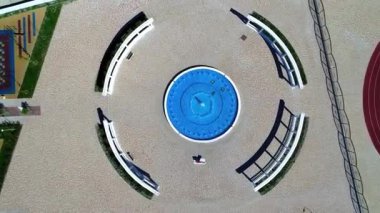 İnsansız hava aracı yavaşça mavi bir çeşmenin üzerinde su sıçratarak yükselir. Sütunlar fıskiyenin etrafında yarım daire şeklinde duruyor.