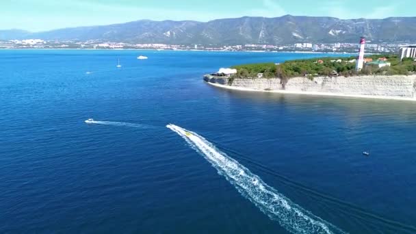 O resort de Gelendzhik, Cabo Gordo. Iates, jet ski puxa uma banana com passageiros no fundo do farol Gelendzhik e as montanhas do Cáucaso. Rochas altas e a cúpula de um clube noturno — Vídeo de Stock