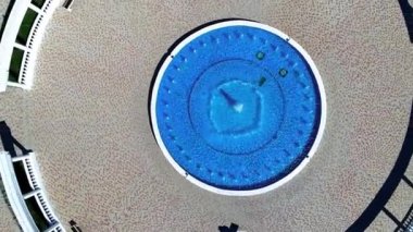 İnsansız hava aracı yavaşça mavi bir çeşmenin üzerinde su sıçratarak yükselir. Sütunlar fıskiyenin etrafında yarım daire şeklinde duruyor.