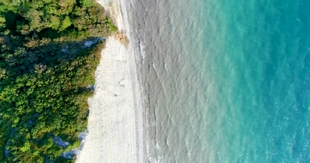 Volando sobre rocas altas y precipitadas cubiertas de bosque. Una pequeña playa de guijarros en el fondo. Esmeralda mar transparente, olas pequeñas — Vídeo de stock