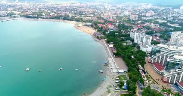 Gelendzhik tatil köyünün merkez plajı kuş bakışı manzaralı. Sıralar dolusu güneş şezlongları ve kumdaki şemsiyeler. İnsanlar yüzer ve güneşlenir. Zümrüt suyu. Sahil boyunca bir kiriş seti var. — Stok video