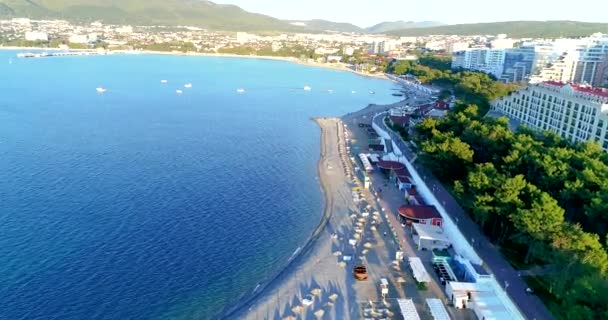 Praia da cidade resort de Gelendzhik, na área de Tolstoy Cape. Gravação de vídeo de um drone. Há fileiras de espreguiçadeiras e guarda-sóis de palha na praia de seixos. Árvore verde. O aterro com — Vídeo de Stock