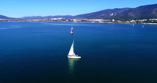 Gelendzhik Körfezi boyunca beyaz yelkenli altında büyük beyaz bir yat, dağlar, Gelendzhik tatil köyü, evler ve plajlara karşı. Dağ olan büyük bir su kütlesi. — Stok video