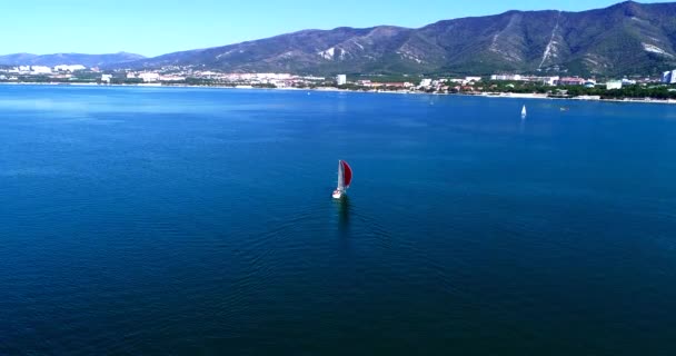 Un hermoso yate blanco grande bajo una vela roja navega lentamente a lo largo de la bahía de Gelendzhik contra el telón de fondo de las montañas del complejo de Gelendzhik. Un gran cuerpo de agua — Vídeos de Stock