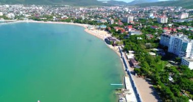 Gelendzhik Tatil Köyü. Kuşbakışı kumsalın üzerinden uçuyor. Çakıl taşlı bir sahil, sıra sıra güneş şemsiyeleri ve güneş şezlongları. İnsanlar yüzer ve güneşlenir. Balüstradlı toprak seti. Geniş bir manzara.
