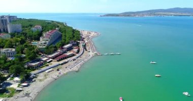 Gelendzhik Tatil Köyü. Kuşbakışı kumsalın üzerinden uçuyor. Çakıl taşlı bir sahil, sıra sıra güneş şemsiyeleri ve güneş şezlongları. İnsanlar yüzer ve güneşlenir. Balüstradlı toprak seti. Geniş bir manzara.
