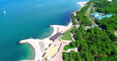 Gelendzhik Tatil Köyü. Kuşbakışı kumsalın üzerinden uçuyor. Çakıl taşlı bir sahil, sıra sıra güneş şemsiyeleri ve güneş şezlongları. İnsanlar yüzer ve güneşlenir. Balüstradlı toprak seti. Geniş bir manzara.