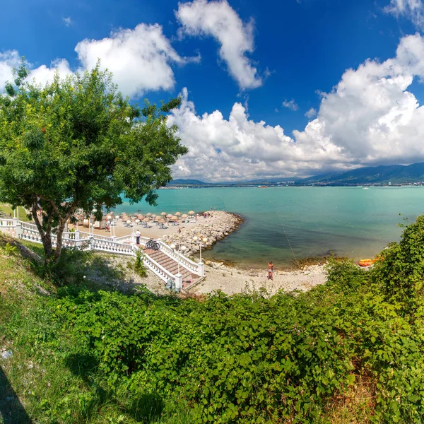Gelendzhik Tatil Köyü. Balyozlu bir merdiven ve kiremit plajına fenerler iniyor. Kumsalda güneşten gelen taşlardan ve şemsiyelerden yapılmış bir iskele var.. 