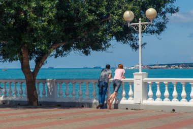 Genç bir çift - bir erkek ve bir kadın, yeşil bir ağacın altında Gelendzhik tatil köyünün setinde duruyorlar ve denize bakıyorlar.. 