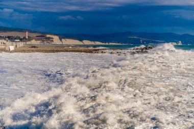 Fırtınada, kar beyazı köpüklü dalgalar Gelendzhik çakıl taşı plajına düşer. Arka planda Gelendzhik deniz feneri ve kayalar
