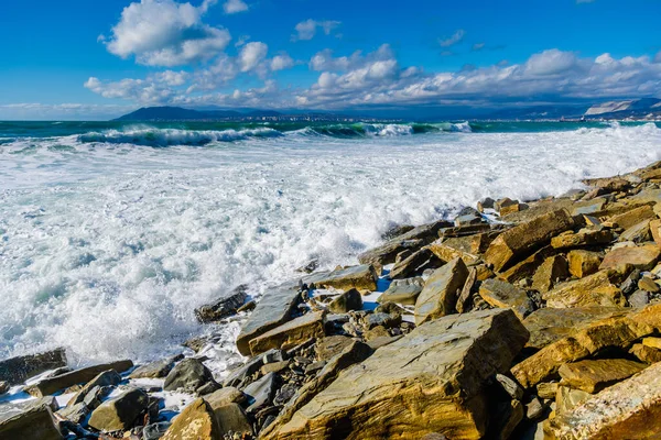 Storm in Tsemesskaya Bay. Turkoois golven met prachtig sneeuwwit schuim vallen op Cape Doob. Er liggen grote geelachtige rotsblokken aan de kust. Aan de horizon Novorossiysk en de bergen — Stockfoto