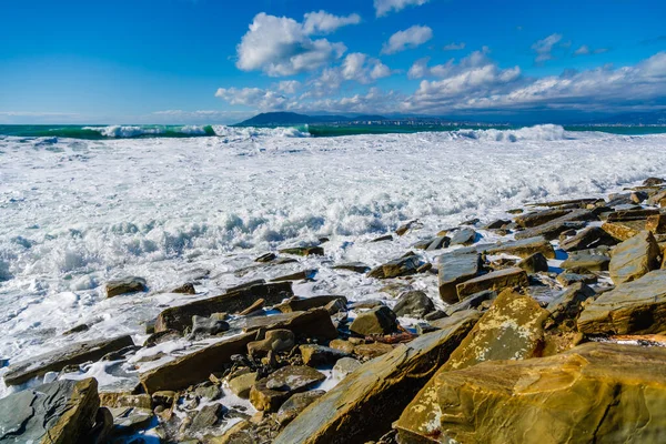 Tempestade em Tsemesskaya Bay. Ondas azul-turquesa com bela espuma branca de neve caem no Cabo Doob. Há grandes pedaços amarelados de rocha na costa. No horizonte Novorossiysk e as montanhas — Fotografia de Stock