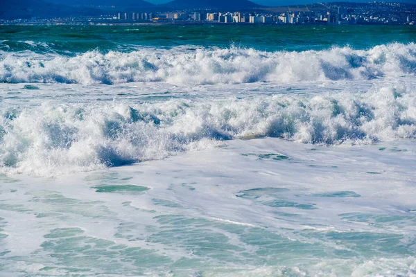 Hermosas olas de tormenta esmeralda en espuma blanca sobre el fondo del monte Myshako y la ciudad de Novorossiysk. Una vista hermosa y peligrosa. Tonos de color azul-verde —  Fotos de Stock