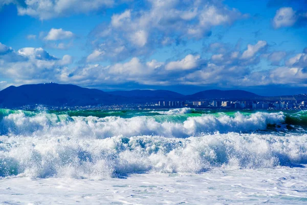 Belas ondas de tempestade de esmeralda em espuma branca contra o fundo do monte Myshako e da cidade de Novorossiysk. Uma vista linda e perigosa. Tons de cor azul-verde — Fotografia de Stock