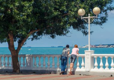 Genç bir çift - bir erkek ve bir kadın, yeşil bir ağacın altında Gelendzhik tatil köyünün setinde duruyorlar ve denize bakıyorlar.. 