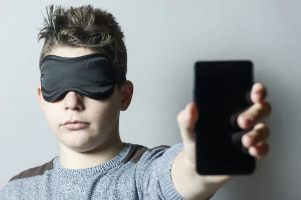 Menino sem sono com máscara de sono segurando e mostrando telefone inteligente — Fotografia de Stock