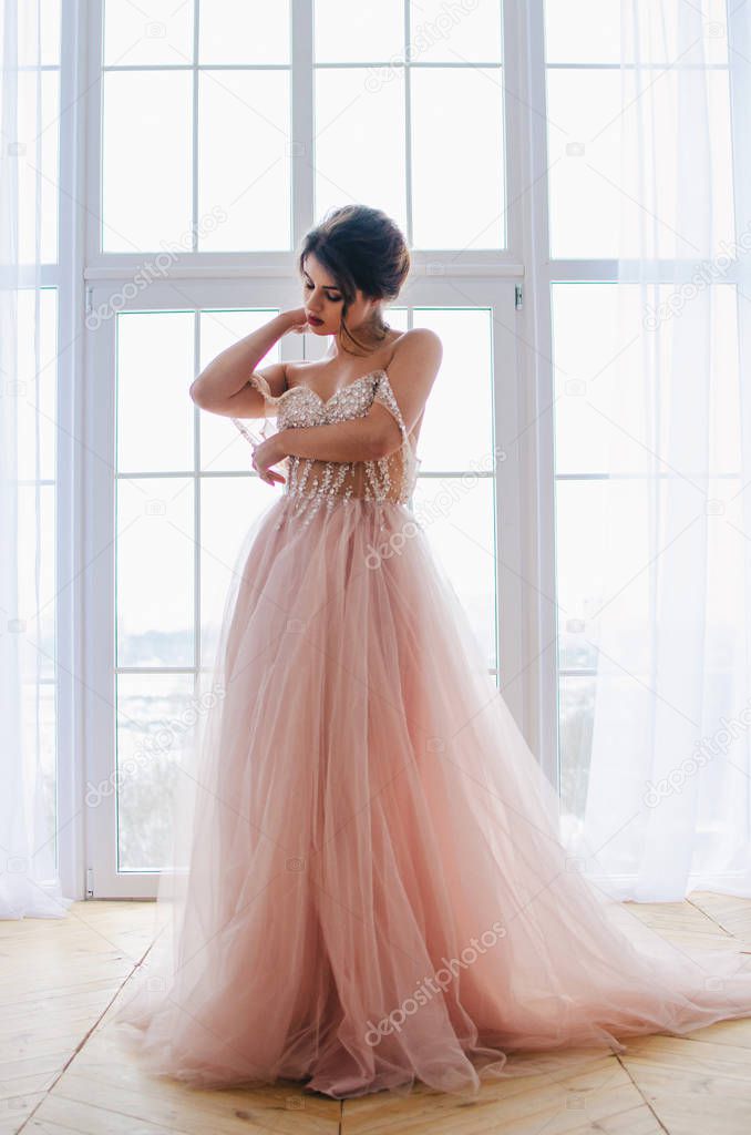 Beautiful girl in a luxurious pink dress is standing near the window