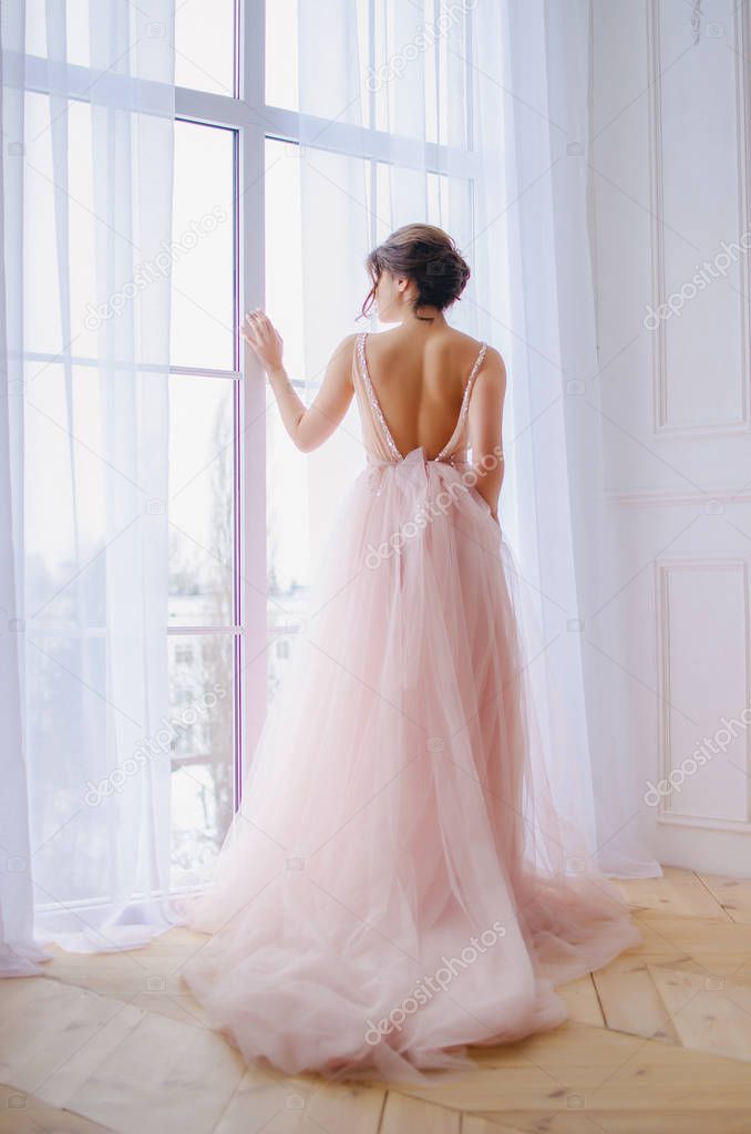 Beautiful girl in a luxurious pink dress is standing near the window