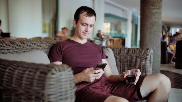 Молодий чоловік друкує на мобільному телефоні і п'є віскі, сидячи на дивані в готелі — стокове відео