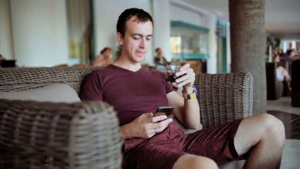 若い男型携帯電話とウイスキーを飲みながらホテルのソファーに座っていた — ストック動画