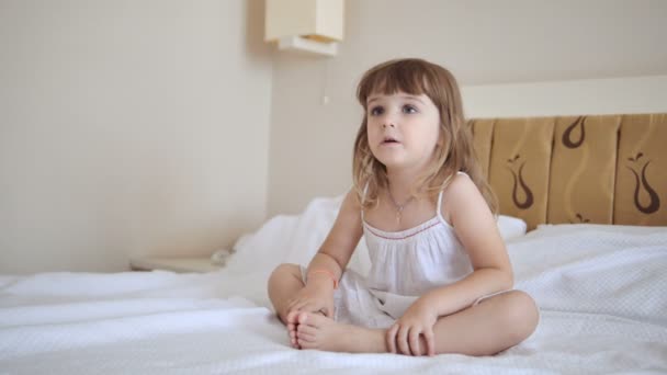 Entzückendes Kind im weißen Kleid vor dem Fernseher auf dem Bett — Stockvideo