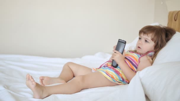 Чарівна дитина в барвистій сукні дивиться телевізор на ліжко — стокове відео