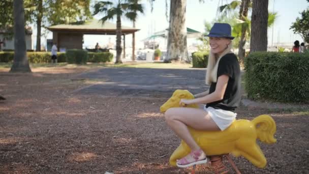 Молодая красивая женщина катается на игрушечной лошади в парке детская площадка — стоковое видео