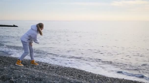 Mujer joven con chaqueta lanzando guijarros al océano, olas espumosas salpicando en tierra — Vídeo de stock