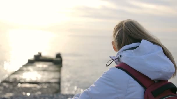 Ung vacker kvinna i jacka med ryggsäck sitter nära havet i solnedgången — Stockvideo