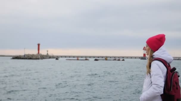 Schöne junge Frau in Jacke und Rucksack spazieren auf der Seebrücke am Hafen. Attraktive Touristin besucht Hafen — Stockvideo