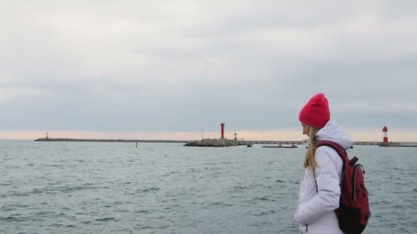 Mooie jonge vrouw in jas met rugzak lopen op pier in harbor. Aantrekkelijke vrouwelijke toeristische bezoekt haven — Stockvideo