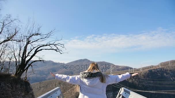 Jovem mulher alegre levantando os braços no skybridge em frente ao belo topo da montanha sobre o céu dourado do pôr do sol no cânion — Vídeo de Stock