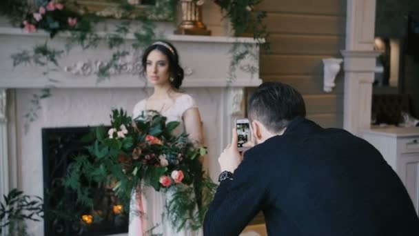 Фотограф снимает невесту с букетом по сотовому телефону. Камин на заднем плане — стоковое видео