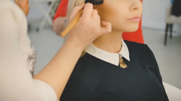 Sarı uzun saçlı güzel genç şehvetli model yüzünde makyaj uygulamak profesyonel makyöz görüntüsünü kapatın. Güzellik ve moda kavramı — Stok video