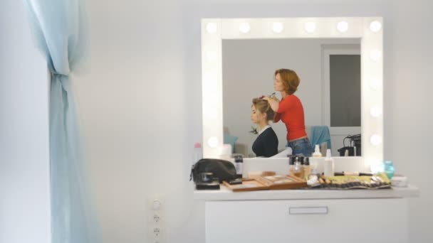 Reflexión a través del espejo - Peluquería femenina de salón de belleza de lujo está de pie y trabajando con la joven modelo rubia — Vídeos de Stock